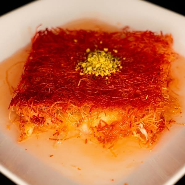 A square kunefe dessert, topped with crushed pistachios, sits in syrup on a white plate.