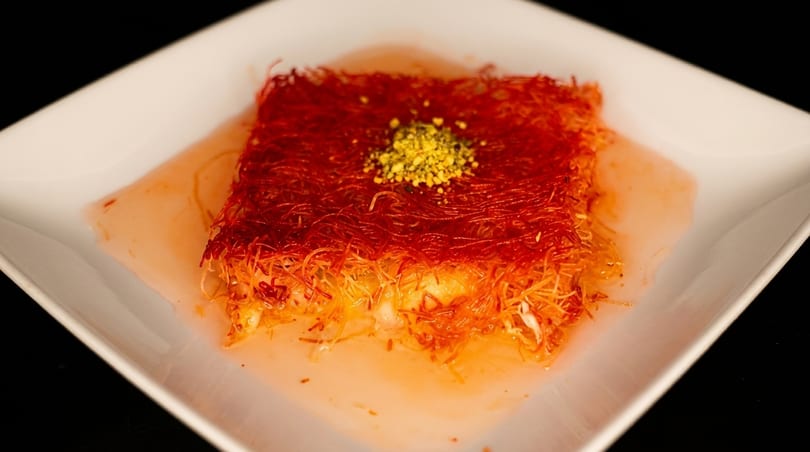A square kunefe dessert, topped with crushed pistachios and soaked in sweet syrup, on a white plate.