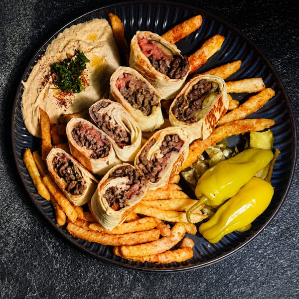 Platter of beef shawarma wraps, french fries, hummus, and pickled peppers on a dark plate.