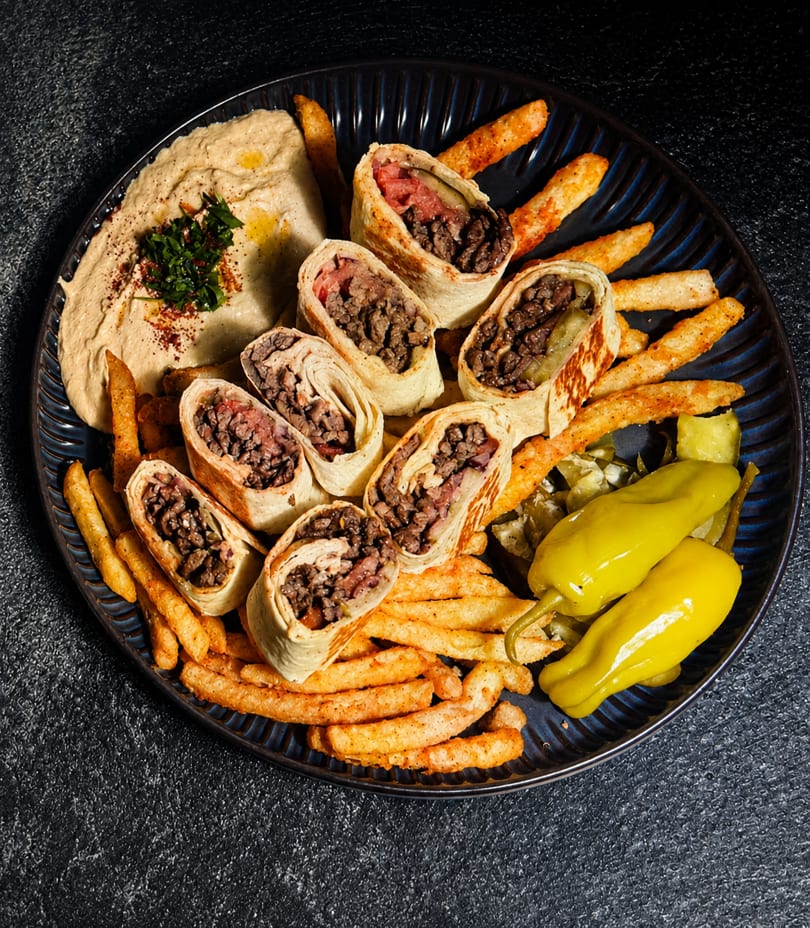 Platter of beef shawarma wraps, french fries, hummus, and pickled peppers on a dark plate.