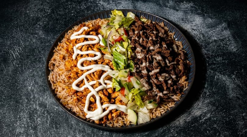 A platter of rice topped with chicken, beef, salad, and white sauce, on a dark background.