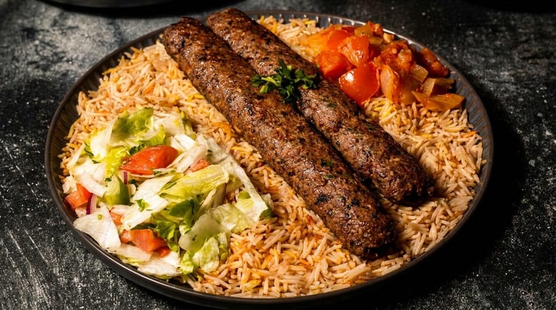 Two grilled meat skewers on a bed of rice, with a side of fresh salad and cooked vegetables.