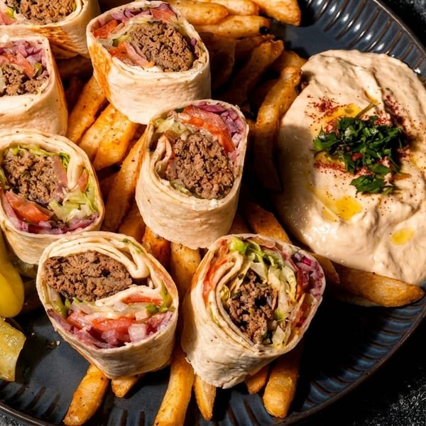 Beef shawarma wraps with fries, hummus, and pickled peppers on a dark plate.
