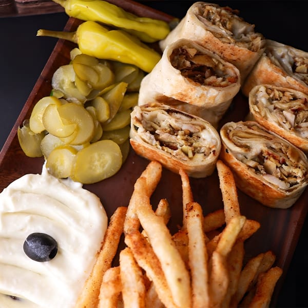 Chicken shawarma wraps, french fries, pickles, and garlic sauce with an olive on a wooden tray.