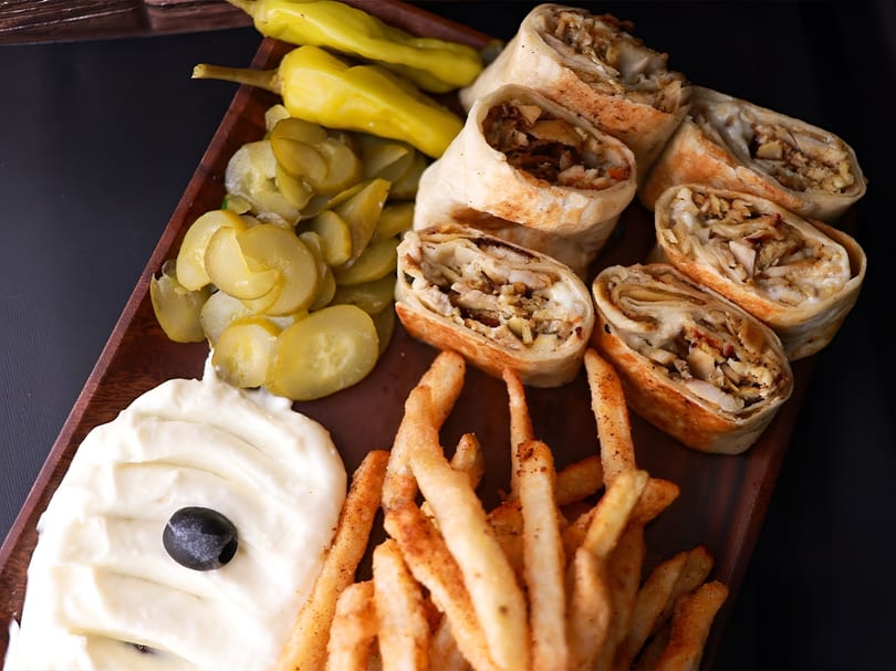 Chicken shawarma wraps, french fries, pickles, and garlic sauce with an olive on a wooden tray.