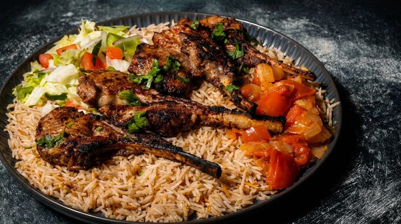 Grilled lamb chops with rice, fresh salad, and stewed tomatoes and onions on a dark plate.